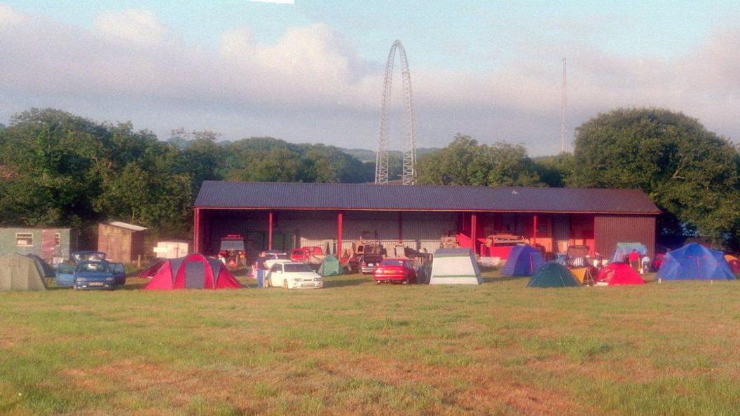 Lots of tents can be seen in the foreground pitched in a field. Behind them is a  storage facility which has farming equipment sitting inside it. In the background there is a pylon as well as several trees.