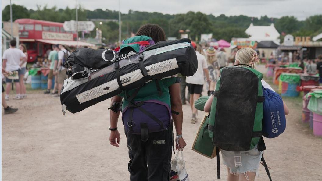 Rear shot of man and woman carrying tents and belongings on their back as they walk along gravel