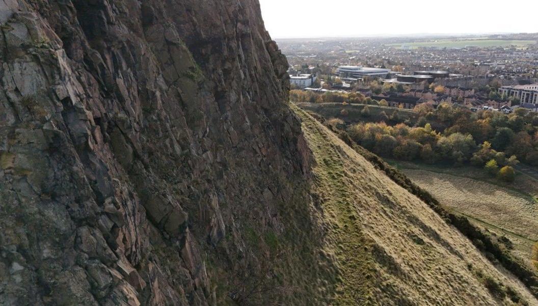 A drone camera view of the overgrown Radical Road path next to the rockface of the Salisbury Crags