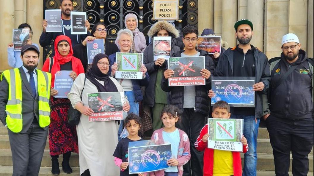 A group of men, women and children protesting with placards in Bradford, asking police to act over the use of fireworks. They are standing on the steps of a grand civic building.