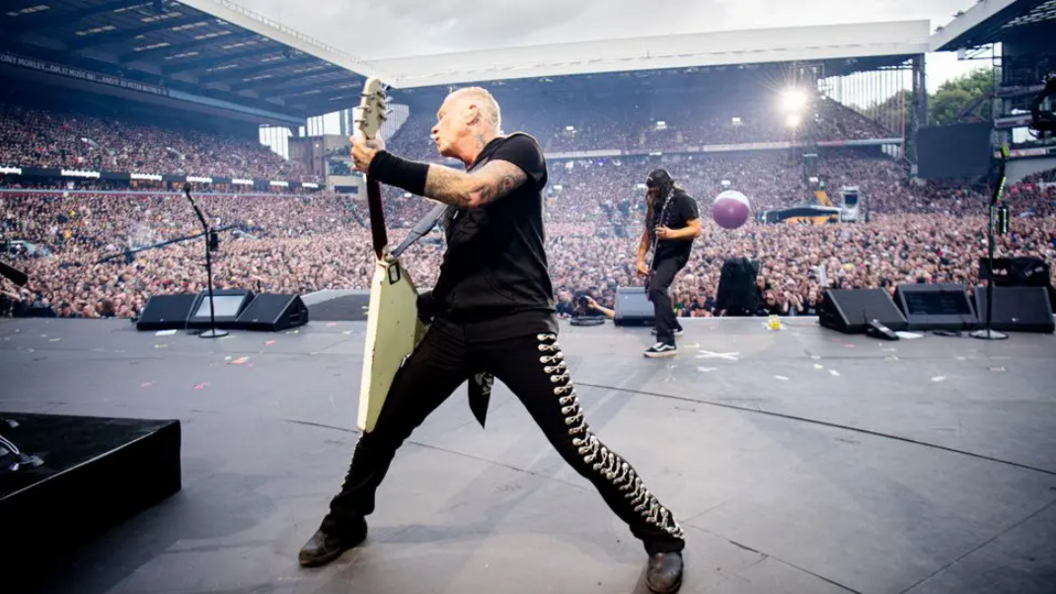James Hetfield, dressed in black with a white guitar and standing with his back to the huge crowd at Villa Park, as he plays a riff and looks over his shoulder. Another guitarist in the band can be seen standing playing close to the edge of the stage.