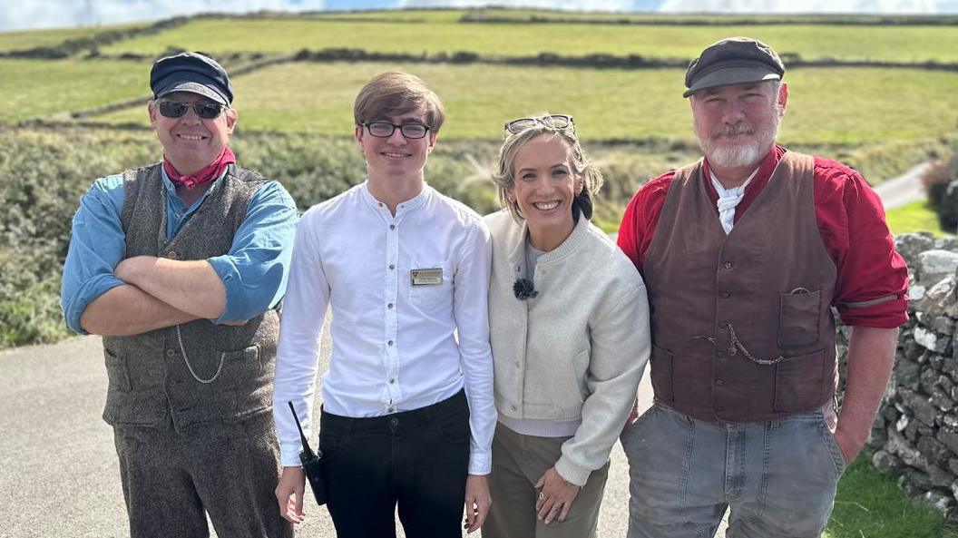 Left to right: Pete Kelly, Lucas Hayhurst, Kay Crewdson, and Tony Kornasiewicz, all standing together smiling against a backdrop of rolling green fields. Both Pete and Tony are in shirts and waist coats and flat caps. Lucas is wearing a white shirt, and Kay has a cream jacket on.