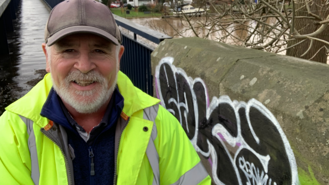 A man with a white beard and grey baseball cap is stood next to the River Severn. He is wearing a blue zip top with a yellow high vis jacket. There is a tree and some graffiti on the wall behind him and a section of the river can be seen.