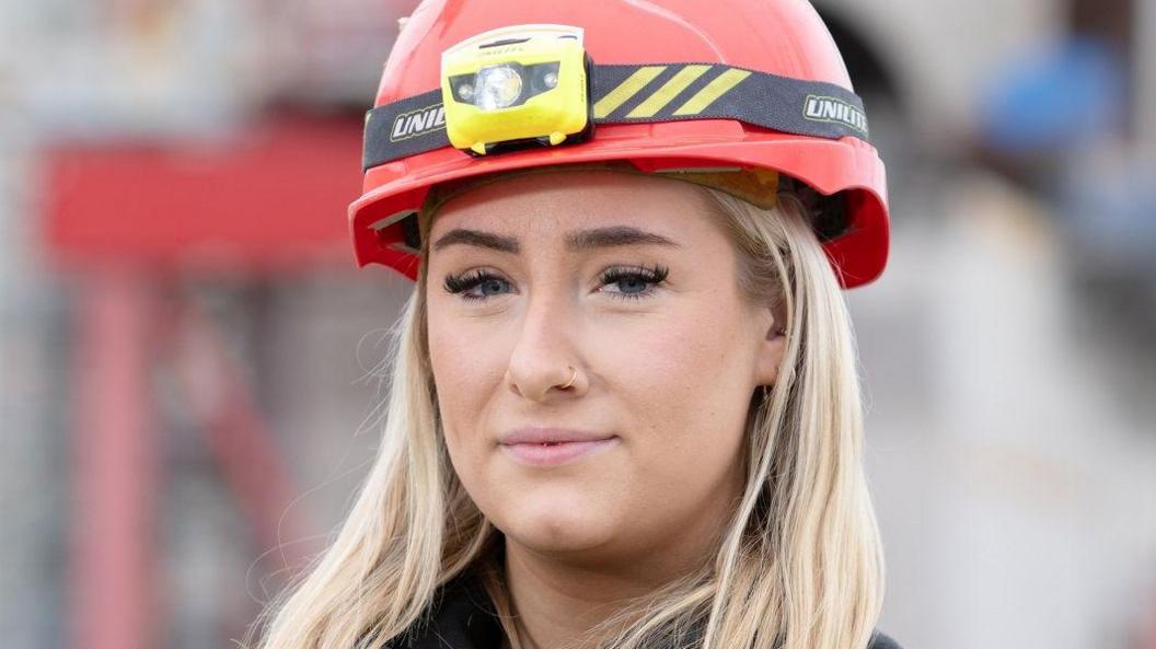 Megan Ellicott has blonde hair and wears a red hard hat with a yellow flash light.