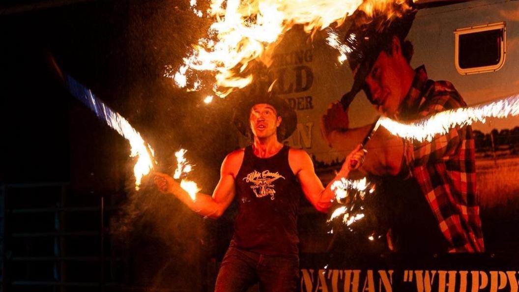A man stands holding two long and burning stock whips. He wears a large hat and a vest and stands in front of a sign with his face on it.