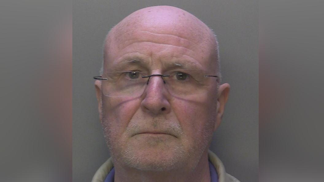 A bald man with glasses and grey stubble stares into the camera for a police mugshot.