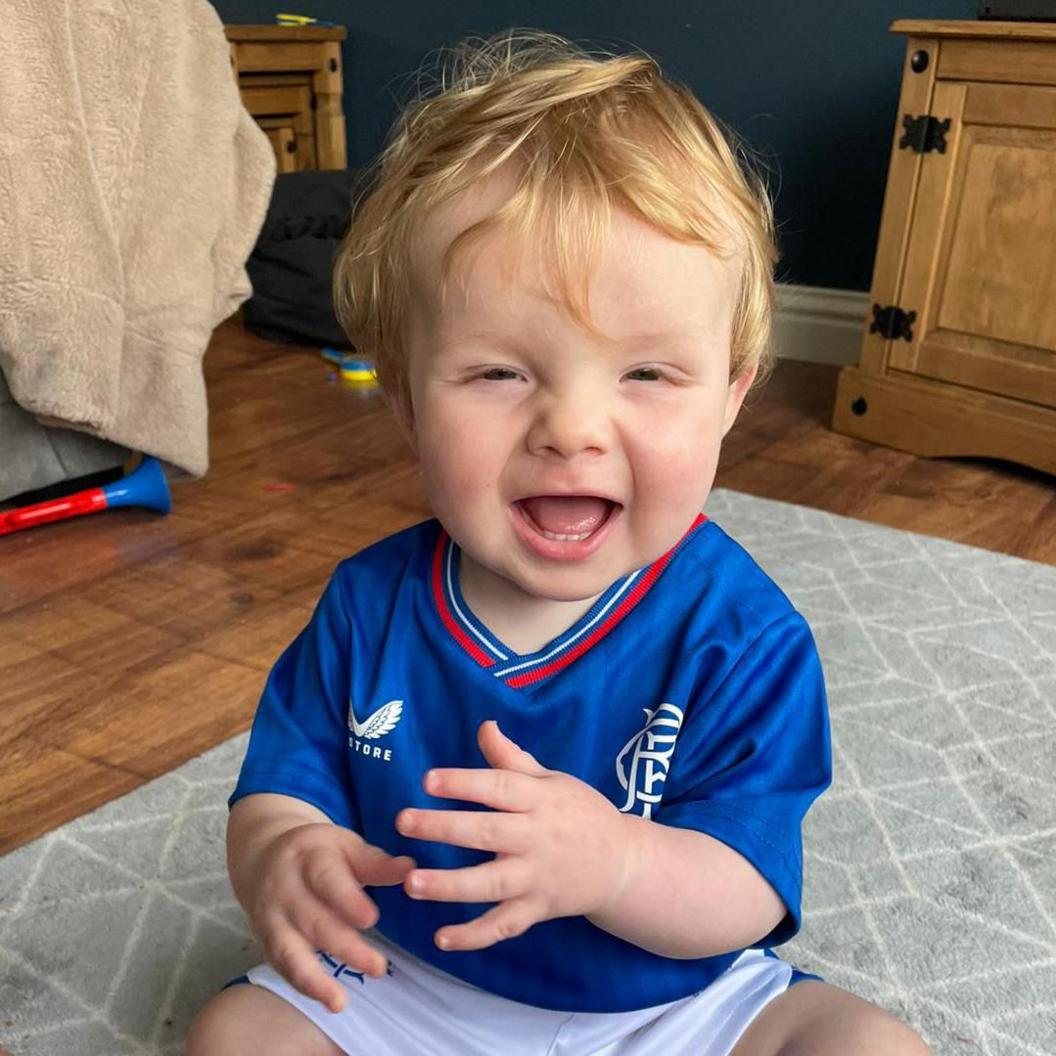 A laughing baby sitting on the floor. Toys in the background.