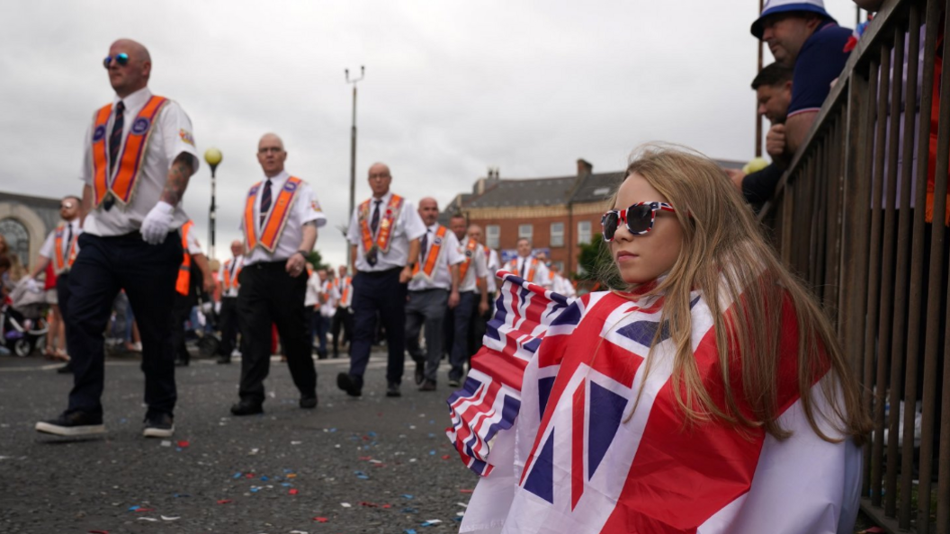 Young girl wrapped in British flag and wearing union jack sunglasses watches Orangemen parade in Belfast