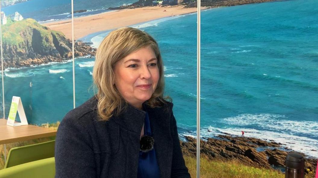 Blond haired lady in a blue coat sat in a cafe with seaside imagery on the walls 