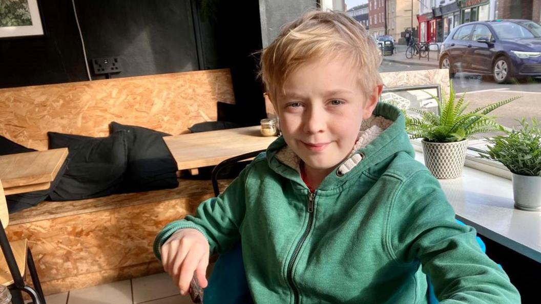 A teenage boy with blonde hair and a green hoodie sat in a cafe, in front of a window with the street behind it and a wooden chair behind him