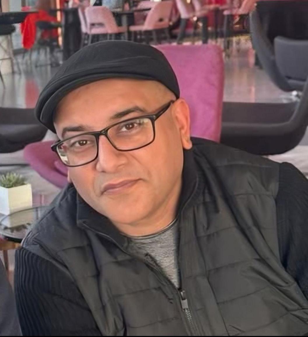 A man with a cap wearing glasses and a coat, smiling at the camera.