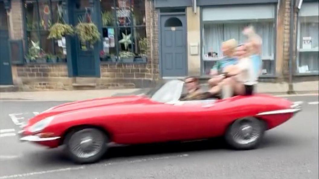 A blurred image of a red e-type Jaguar being driven down a street in Sowerby Bridge, with three people sitting at the back waving.