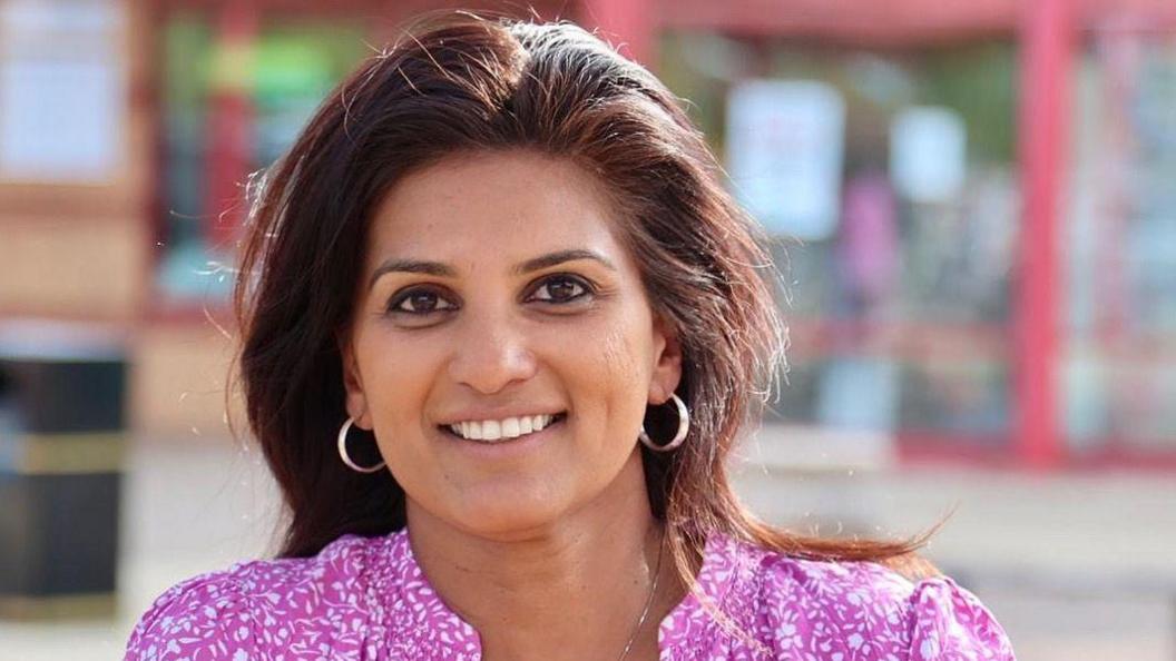 Shazna Muzammil with long dark hair and two large silver earrings smiling at the camera and wearing a pink and white top. The background is blurred but appears to be a shop front with notices attached.