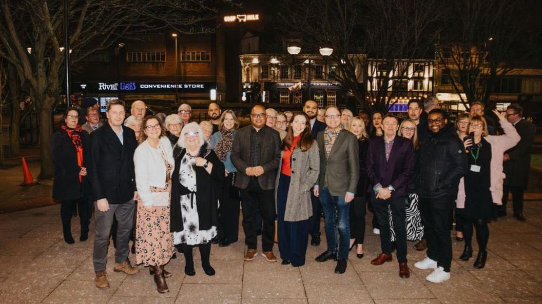 A couple dozen people stand in front of the camera in Derby city centre at night. A illuminated sign which reads COOP MILK  is in the background. 