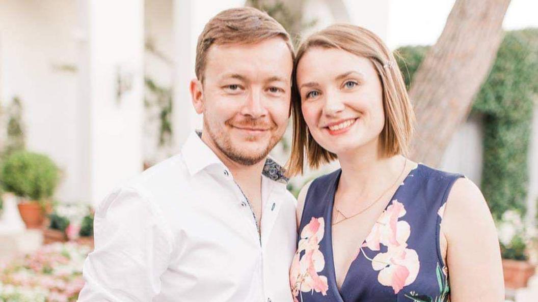 Ricky Skinner and his wife standing next to each other. He is wearing a white shirt and she is wearing a blue dress.