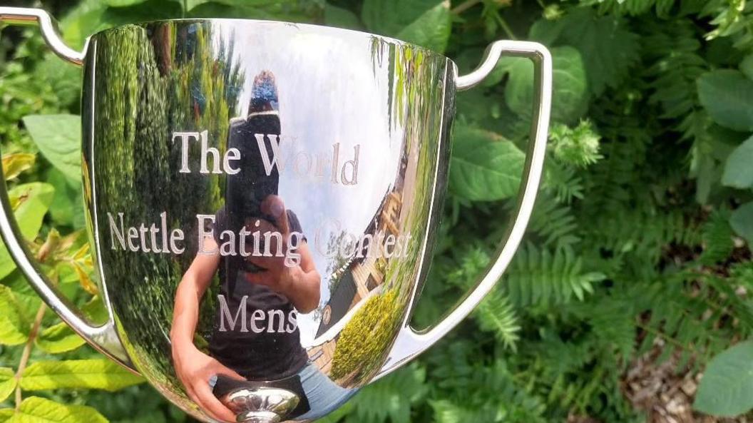 A silver-coloured trophy with handles and engraved with the words, the world nettle eating contest men's. A man holding a camera phone can be seen reflected in the metal.