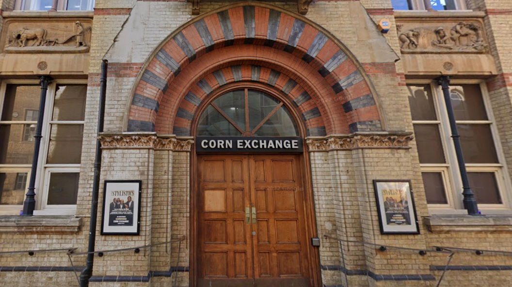 The Google image shows the two big wooden doors with a black sign atop for the Corn Exchange. There are two framed posters either side of the entrance against the cream coloured bricks and railings on both sides of the building leading up to the double doors. There are carvings in the upper level of the building, beneath two sets of windows, showing what looks to be agricultural workers and a horse and plough. 