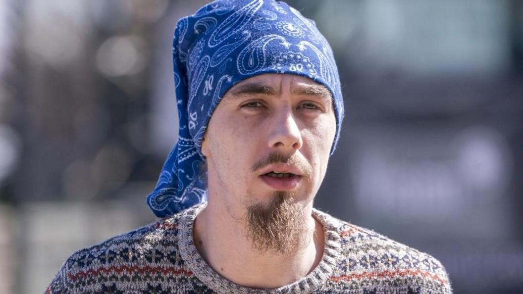 Ashley Darbyshire, 28, with a wispy brown moustache and goatee beard, is pictured wearing a blue bandana tied over his head and a multicoloured woollen jumper. 