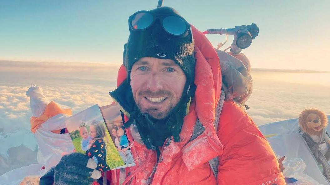 Adam Jones, at the summit of mount everest, holding a photo of his wife and children and smiling.