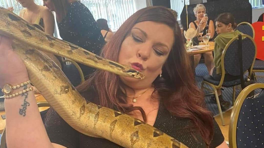 Tara Costellow, Gareth Costellow's wife who helps run the centre, at a social event holding a snake and giving it a kiss. There are people sitting at tables in the background with drinks.