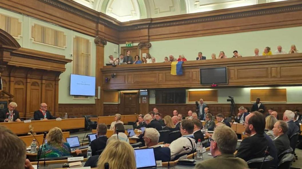 A council chamber full of people with a yellow and blue Ukrainian flag draped over the public gallery balcony