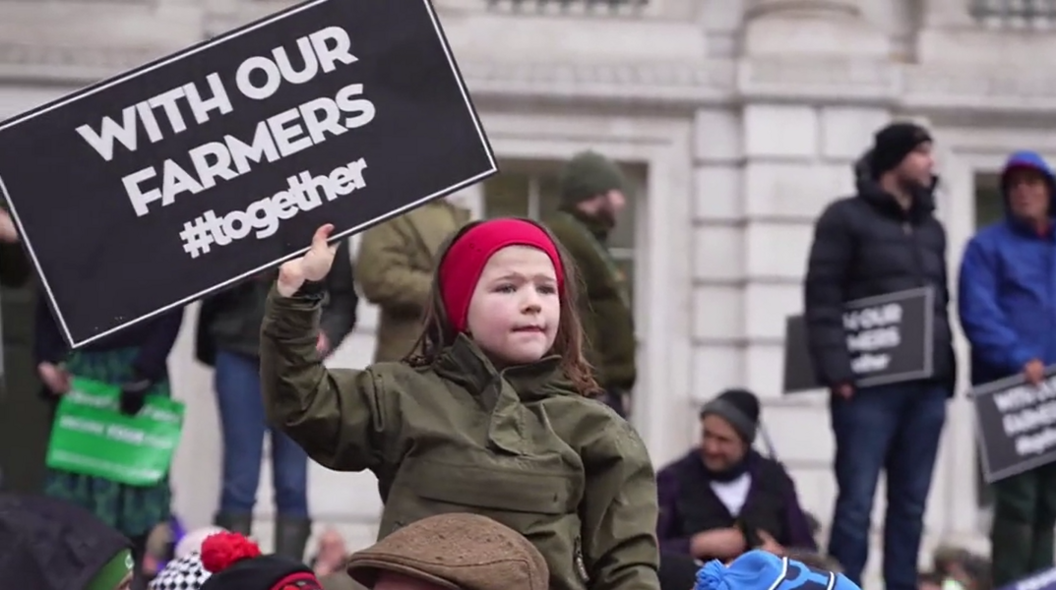 A small child, wearing a green jacket and red hat holding a sign which reads "with our farmers #together" 