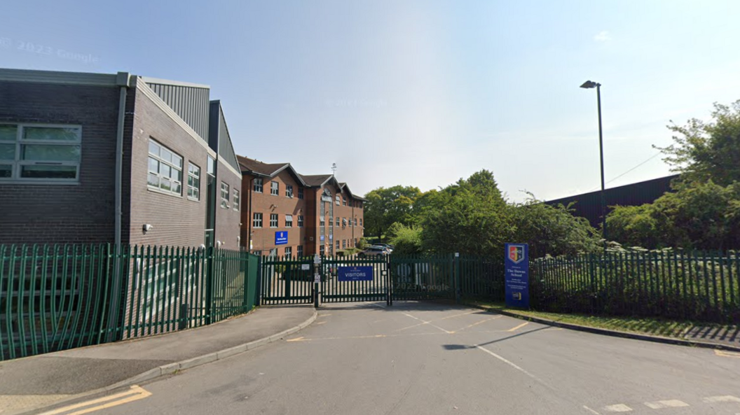 A Google Street View screenshot of the outside of a school. It has dark green railings and double gates closed over the road. A blue sign next to the road says the name of the school and there are several brick buildings behind the railings.