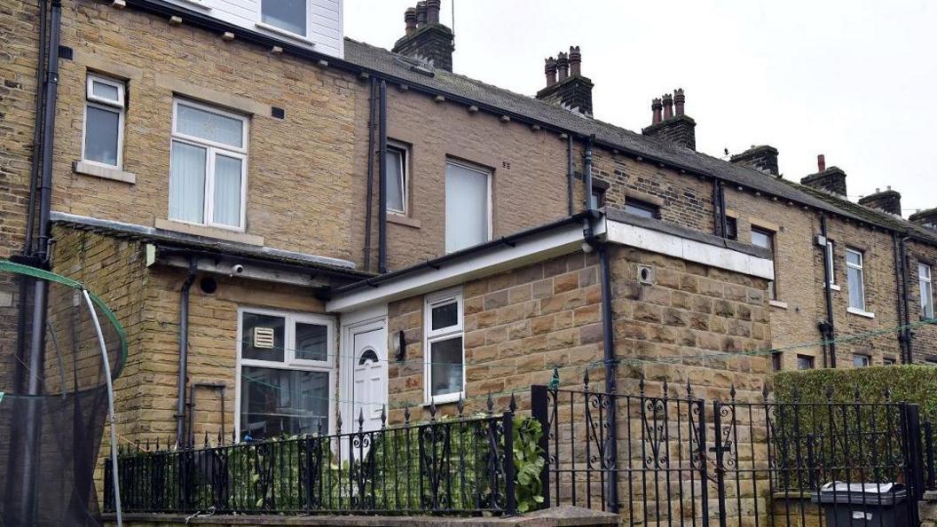 The photograph shows a two-storey property with an extension. A black metal fence surrounds the home and a trampoline can be seen to the left of the property.