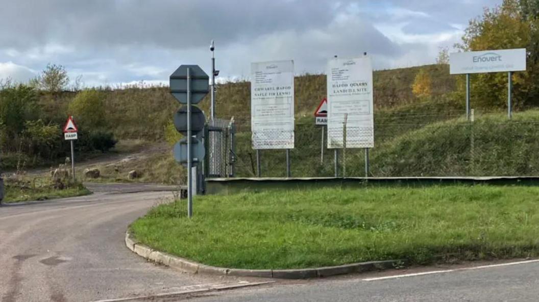A drive leads up to the entrance to the landfill. There is a metal gate and a metal fence running around the entrance with white signs behind with Hafod Quarry written on. A grass verge can be seen behind.