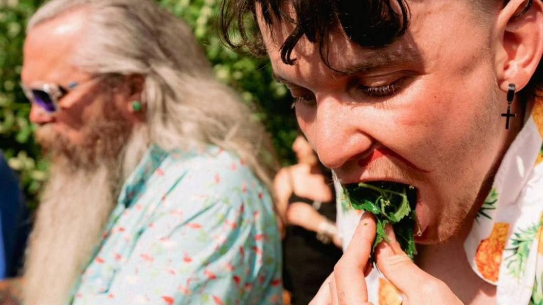 Two men pictured at close range - the man in the foreground has dark hair and is stuffing some nettle leaves into his mouth, while the man in the background has long greying hair and a long greying beard