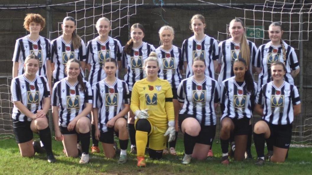 A team photo. The front row are on bended knee, the back row are standing. The women wear black and white striped tops, apart from the goalkeeper in the centre who wears yellow