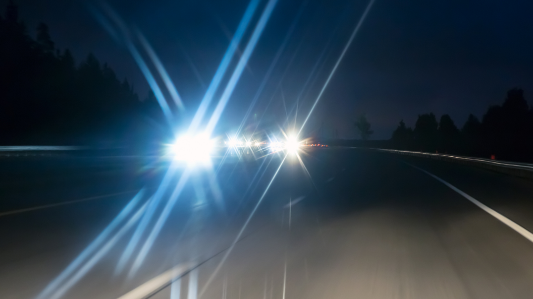 A shot taken at night on a dual carriageway. The carriageway on the right is empty. On the other carriageway, the bright lights of car headlights are flaring in the lens of the camera as they drive towards it. The silhouettes of trees can be seen on either side of the road.