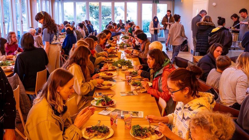 Dozens of people sit on either side of two long tables, eating. There are long windows at the end and left hand side of the room. On the right hand side, people are being served food from a large pot.