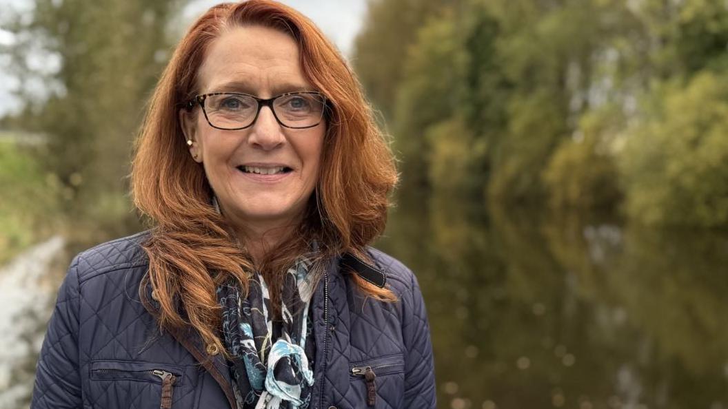 Heather MacLachlan, a woman with red hair and wearing a blue jacket and glasses. There is water in the background.