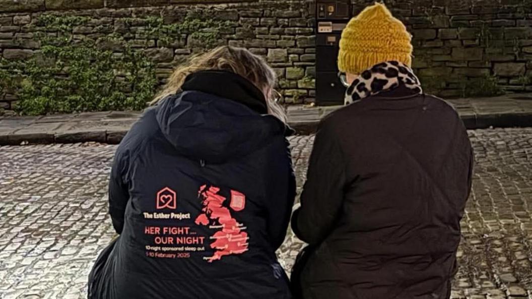 The backs of two women who are sat on a bench in winter coats. Lianne is in a branded coat that says The Esther Project - Her Fight...Our Night.