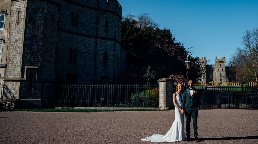 A bride and groom posing for a wedding picture in front of the Windsor Castle, which can be seen in the background. They are both smiling, it is a sunny day.
