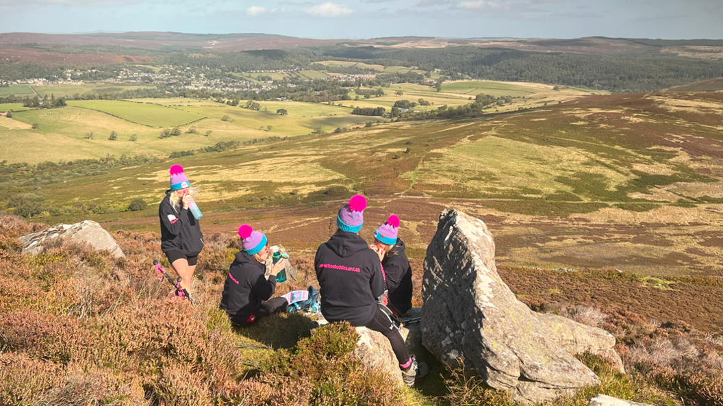 The group of women sat on top of a hill, facing the valley below. They are drinking from water bottles.