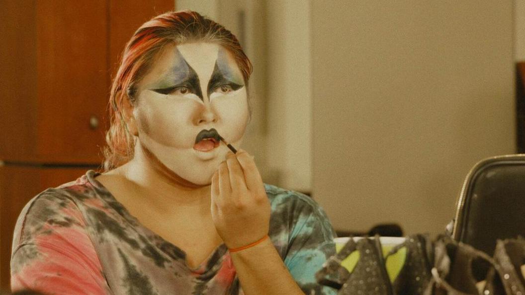 A screengrab from the student documentary Girly showing a person with blonde, long hair putting on face paint. They are wearing a multi-coloured top and are sitting looking at a mirror