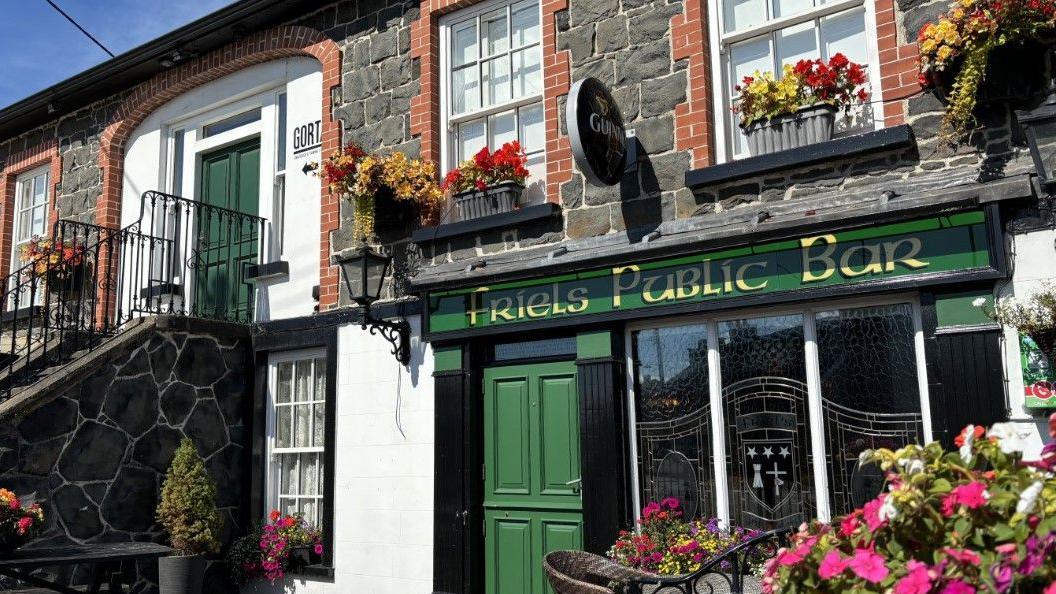 Friel's pub in Swatragh - it is a very traditional Irish bar, with green signage and a door, and a Guinness sign attached. There are flowers in the window boxes and in planters outside.