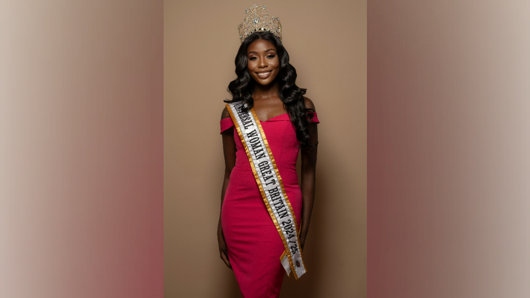 A black woman with long black hair past her shoulders stands facing the camera. She wears a pink-red dress with a long sash which says "Universal Woman Great Britain 2024/25" on it. She has a tiara on her head.