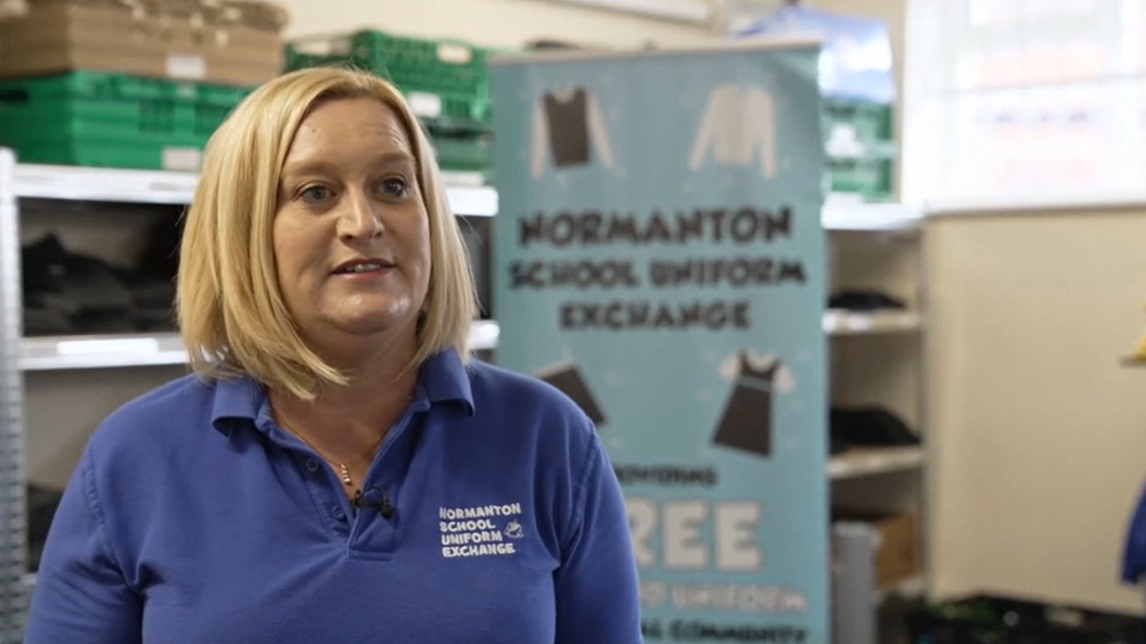 Tracy has blonde shoulder-length hair and is wearing a blue polo t-shirt with the Normanton School Uniform Exchange logo on it. Behind her is school uniform on shelves.