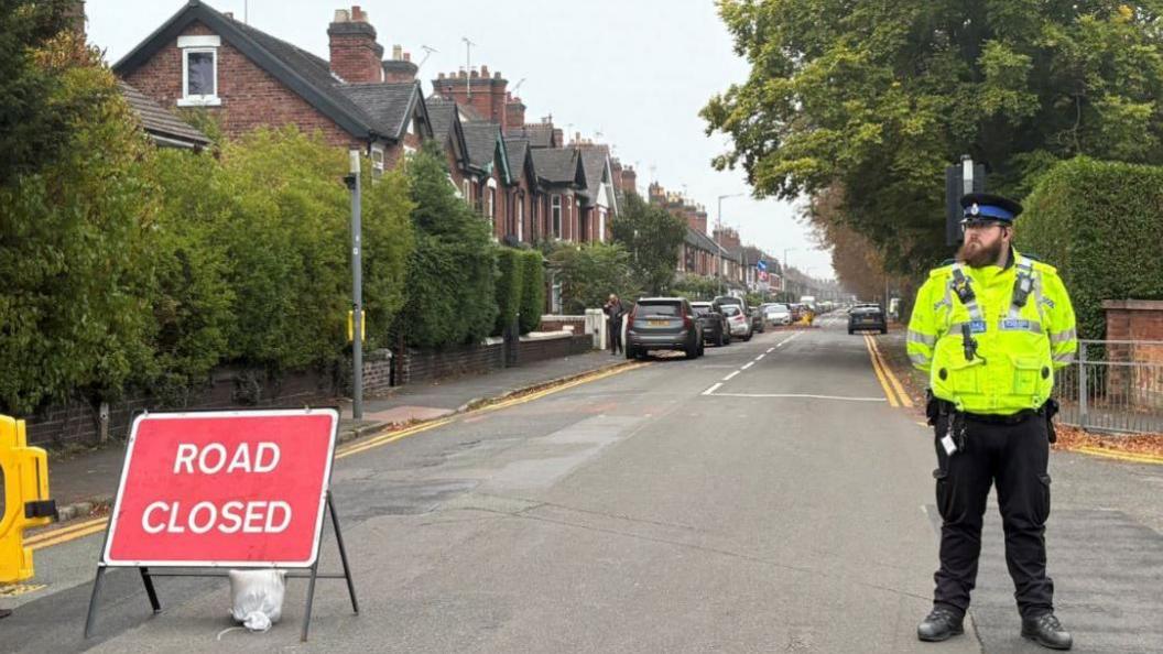 A police officer can be seen standing on the right, with hi-vis jacket and black uniform trousers on. The road where the children were found is behind him, with houses on the left and trees to the right. There is a Road Closed sign on the left of the shot  