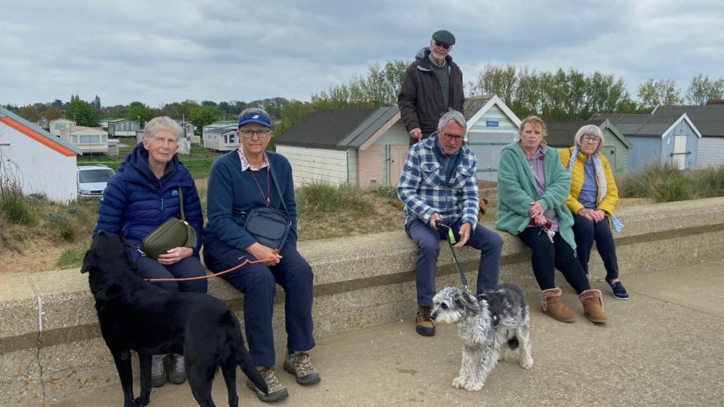 Six people are sitting and standing on concrete in front of a row of beach huts in Heacham. Behind them is a caravan park and a row of trees.