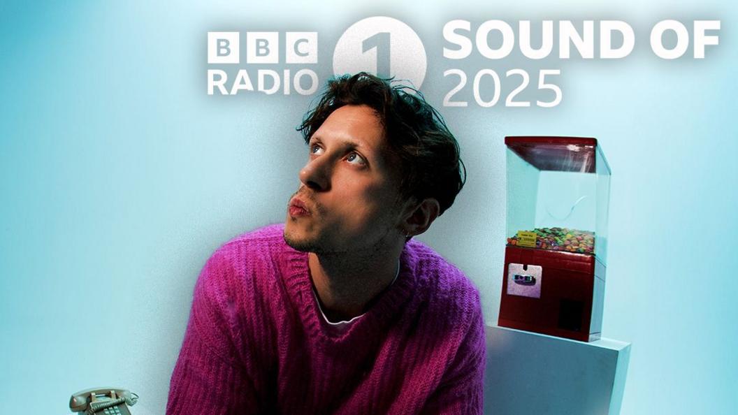 Barry Can't Swim, dressed in a mauve jumper, looks upwards with pursed lips, in a light blue room dressed with a gumball machine and an old-school telephone.