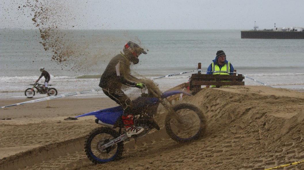 A motocross rider negotiating a sand ramp on the beach course. Sand is flying up from the wheels as they climb the ramp. A safety marshal can be seen behind a barrier wearing high-viz clothing. In the background another rider can be seen on his bike heading in the opposite direction on a different part of the course.