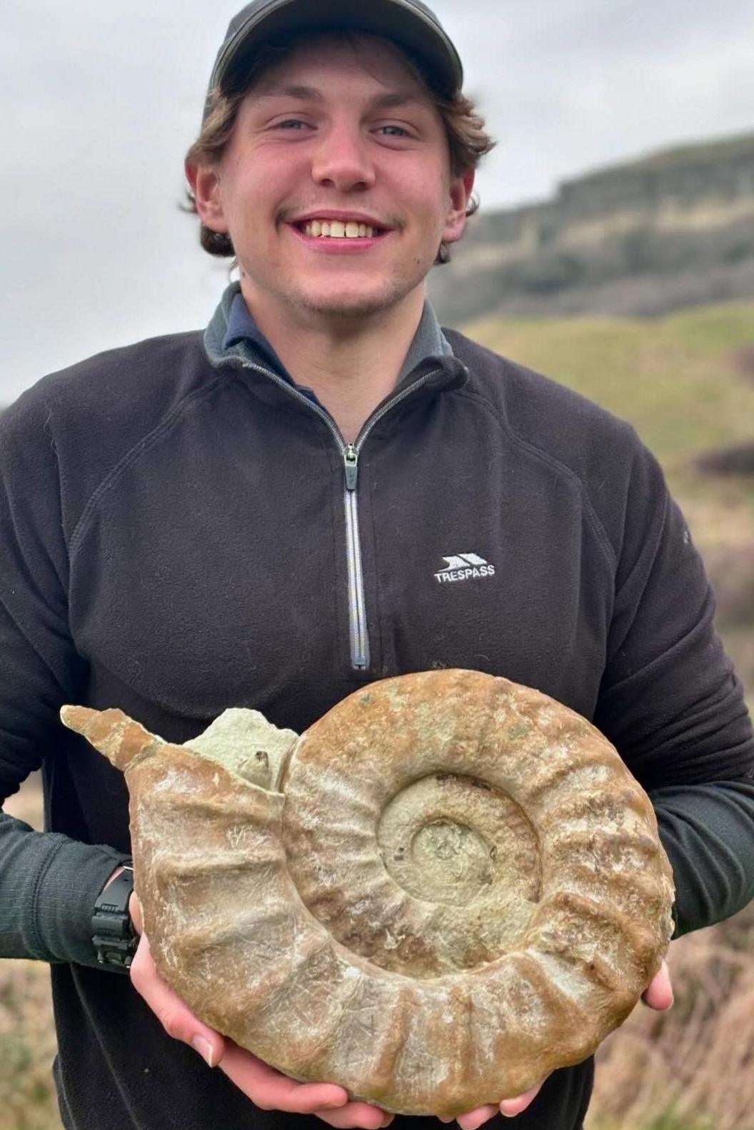 Man in zip-up top with black baseball cap on holding a large fossil.