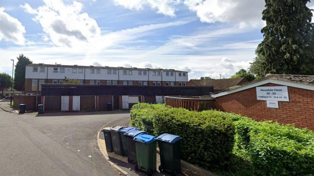 A residential street with a small block of flats and bins lined up by a hedge