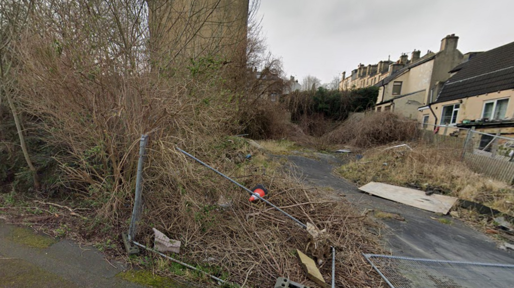 An apparently neglected and overgrown plot of land sited at the end of a residential street. A metal perimeter fence supposedly gating the site off has fallen over and a cone and scrap wood can be seen.