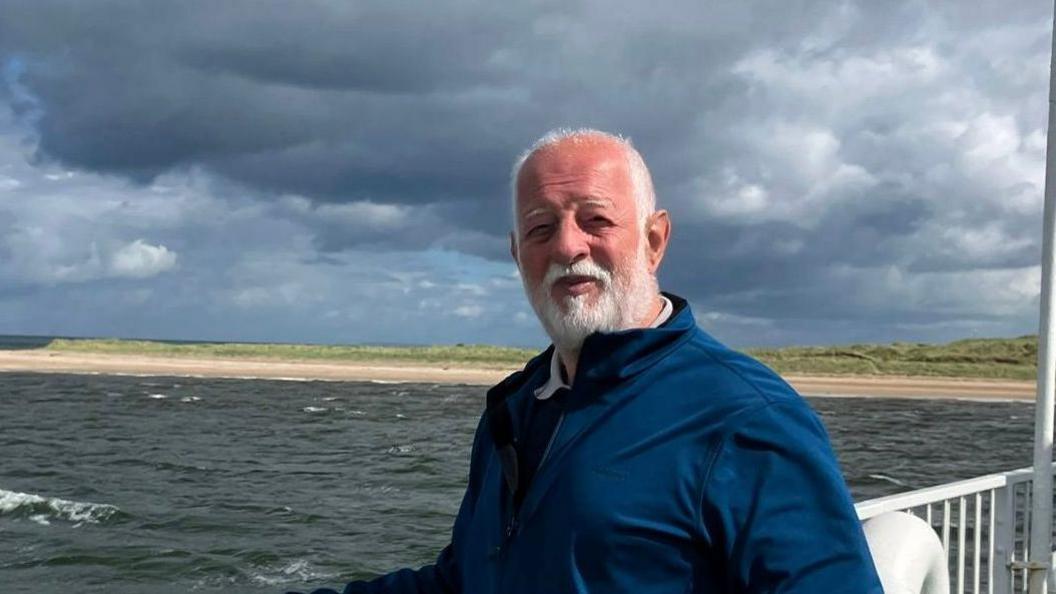 A photo of Barrie, with white hair and stubble, he wears a blue jumper and smiles while being photographed in front of the sea.