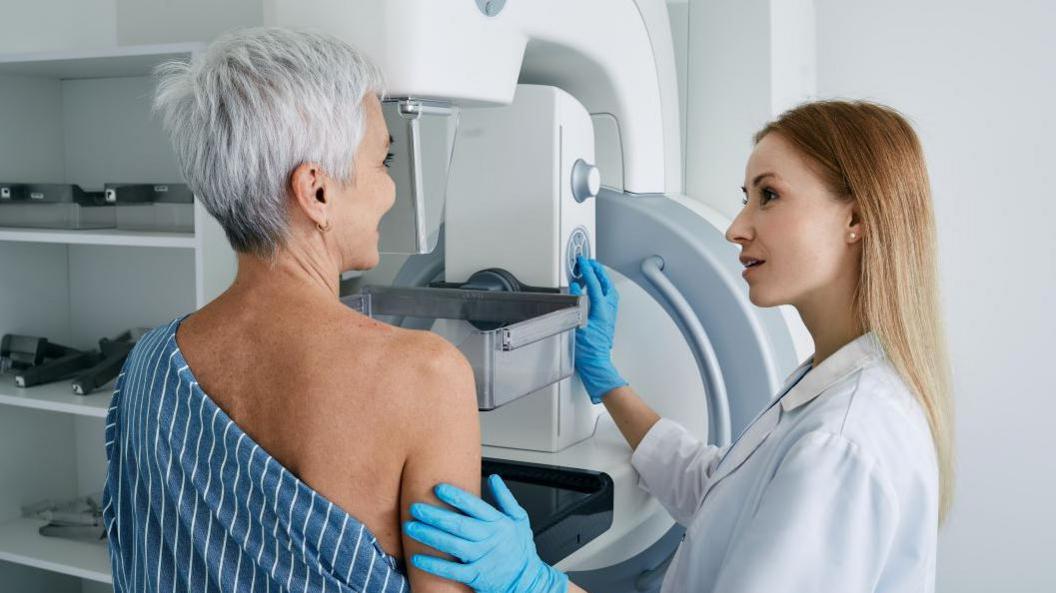 A woman with short grey dyed hair is wearing a blue and white striped gown which is slightly slid down as she receives breast cancer screening. The doctor standing next to her has one hand on her arm and the other on the machine. She has long brown and blonde hair and is wearing a white robe with blue gloves.